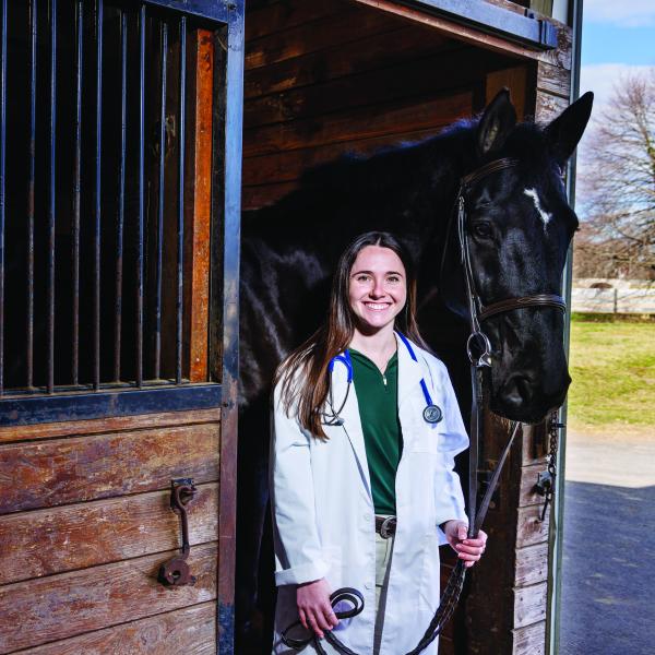 Nina Dellarte, a class of 2021 graduate is in a white coat next to a black horse at the equestrian center.