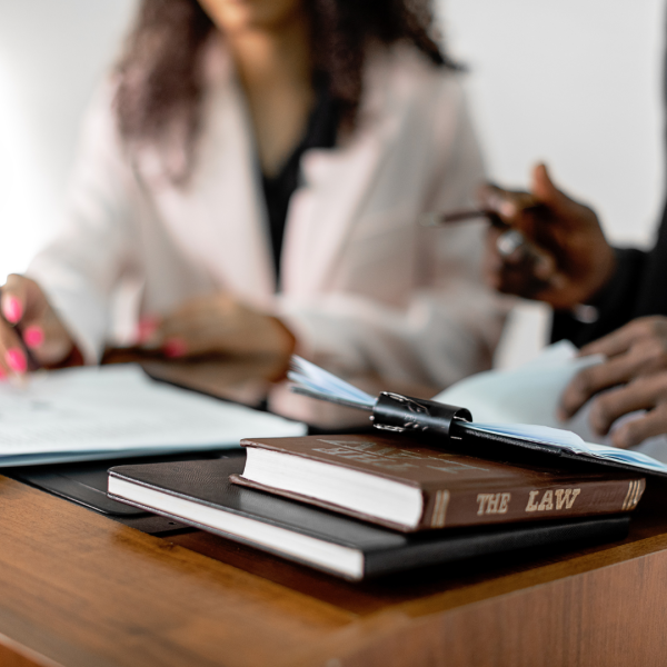 A law textbook is sitting on a desk with two people sitting at the desk writing. 