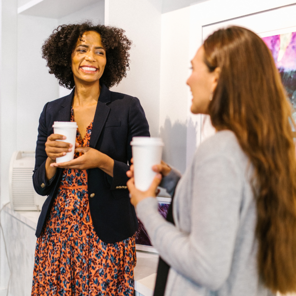 Two female adults dressed professionally are talking with coffee in hand. 