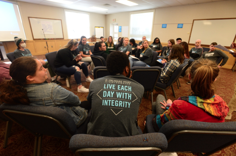 Students are seated in chairs in a circle in a large conference room with no tables. 