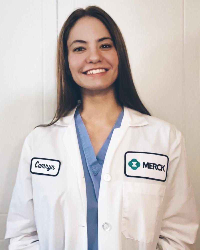 A headshot of Camryn with a white coat with a merck logo on the upper left corner.