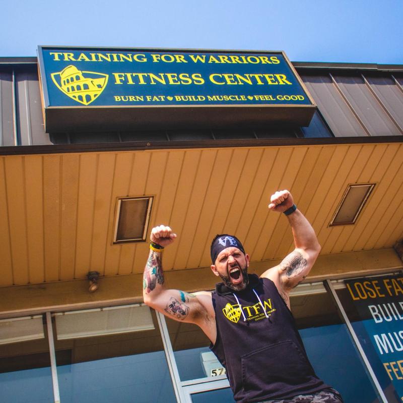 Mike Sobczak poses with arms in the air in front of the training for warriors sign outside. 