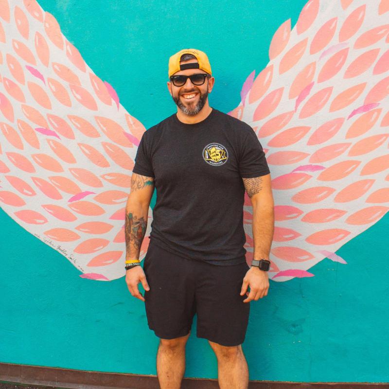 A full body photo of Mike Sobczak '07 wearing a yellow baseball cap, sunglasses, and black shorts and a black shirt.