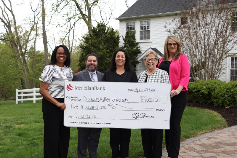 Four DelVal Staff and Two Meridian Bank Staff pose together outdoors with a giant check in hand. 