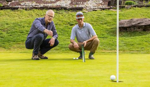 Professor Doug Linde and a Student are on a golf course speaking one-on-one while squatted down on the turf.