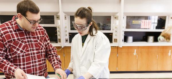 A professor and student wearing safety glasses and looking at a computer screen in a lab.