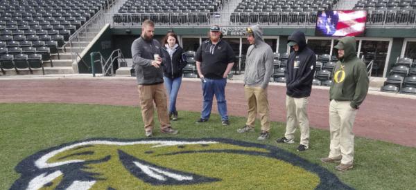 A group of students standing in a stadium on the greens listening to a professional grounds keeper.
