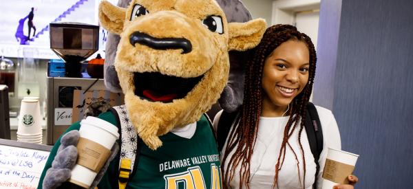 A student holds a Starbucks coffee and stands with the Delaware Valley University mascot who is also holding a Starbucks coffee.