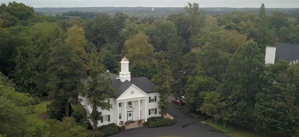 Drone photo of campus with white building in foreground and a vast area of wooded land in the background.