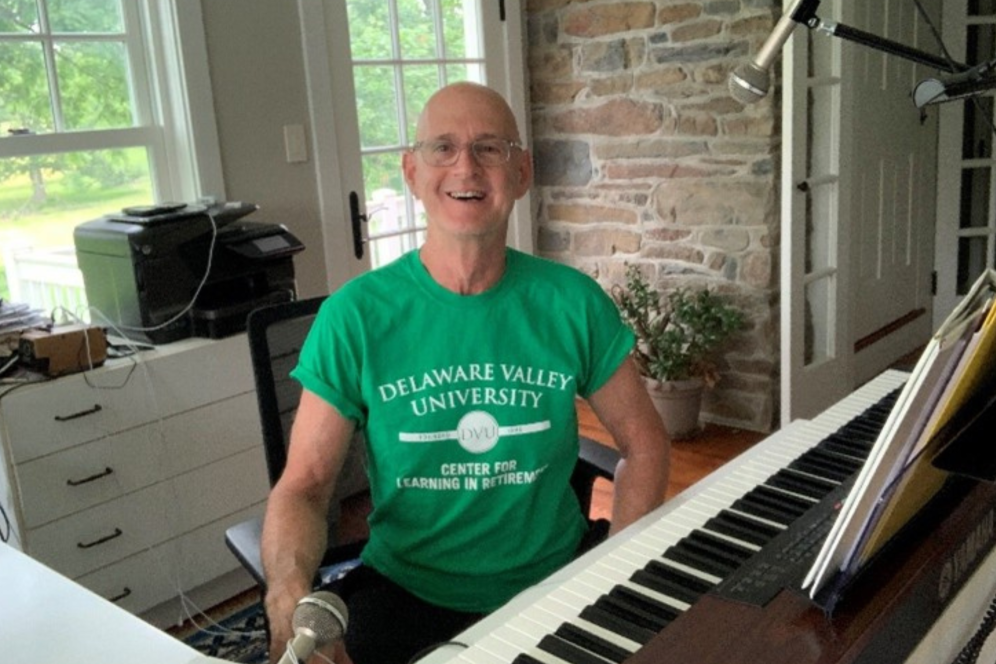 A man wearing a green CLR Shirt is smiling in front of a piano and microphone. 