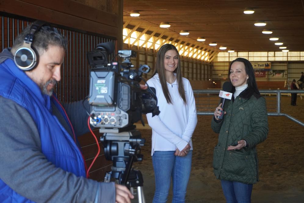 A camera man is filming a student and a reporter in the equestrian center. 