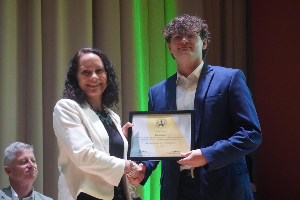 A student is holding an achievement award on stage while shaking Dr. Tanya Casas hand.